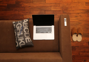 Photo of a laptop, Apple TV remote and a pillow on a couch with brown slippers on the wooden floor